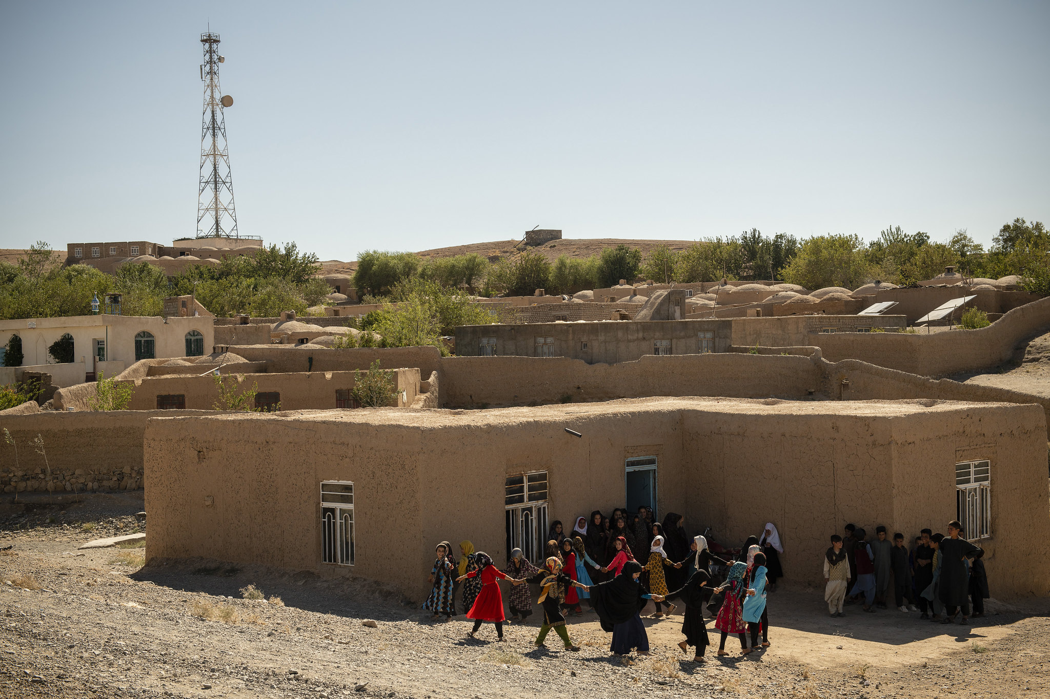 A group of young children hold hands and stand in a circle outside a building in Afghanistan.