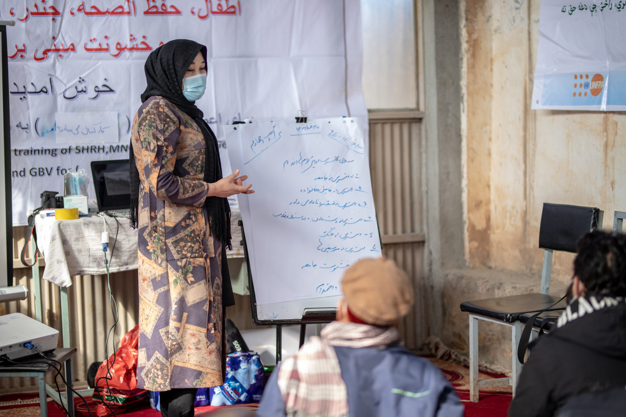 A woman wearing a medical mask, is standing in front of people and leading a health and hygiene training seminar.