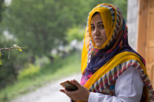 Mufida holds her phone and looks into the camera. She is wearing a colourful headscarf.