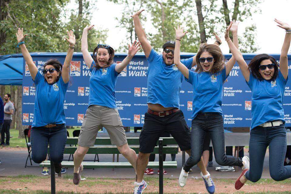 Four young adults are jumping to pose for a photo. They are all wearing blue shirts with an RBC logo. They are in front of a blue backdrop with the World Partnership Walk logo.