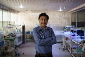 A doctor wearing a blue button-up shirt stands in a NICU ward, smiling with his arms crossed. Behind him are newborn incubators against the walls.