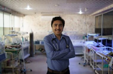 A doctor wearing a blue button-up shirt stands in a NICU ward, smiling with his arms crossed. Behind him are newborn incubators against the walls.