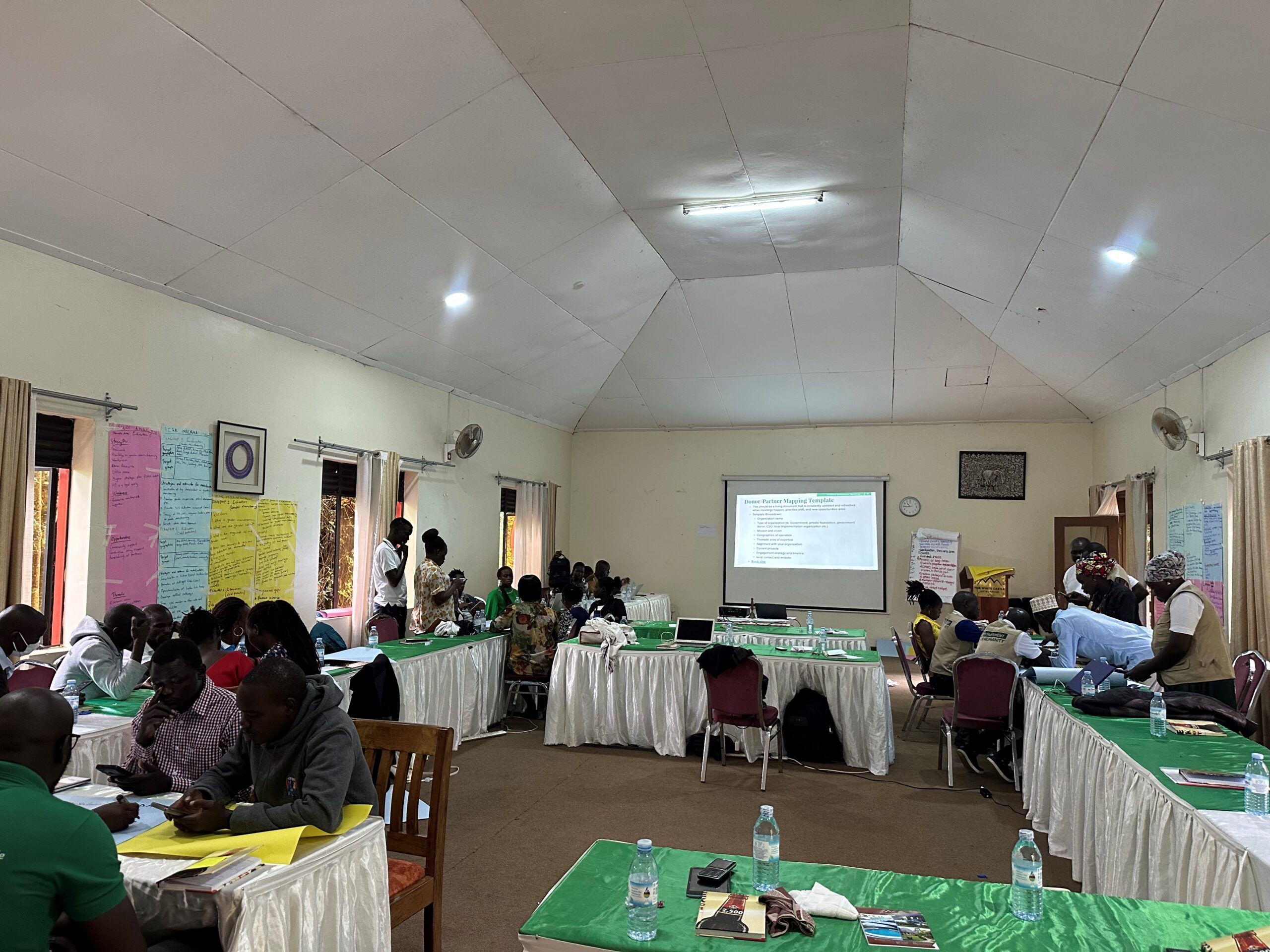 Tables with green tableclothes are set up in a well-lit room with high ceilings..