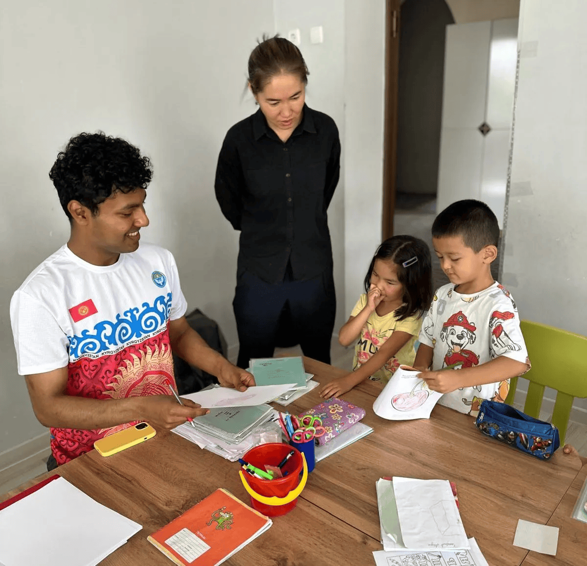 Mahir sits with two kindergarten students at a wooden table. Their teacher is standing nearby.